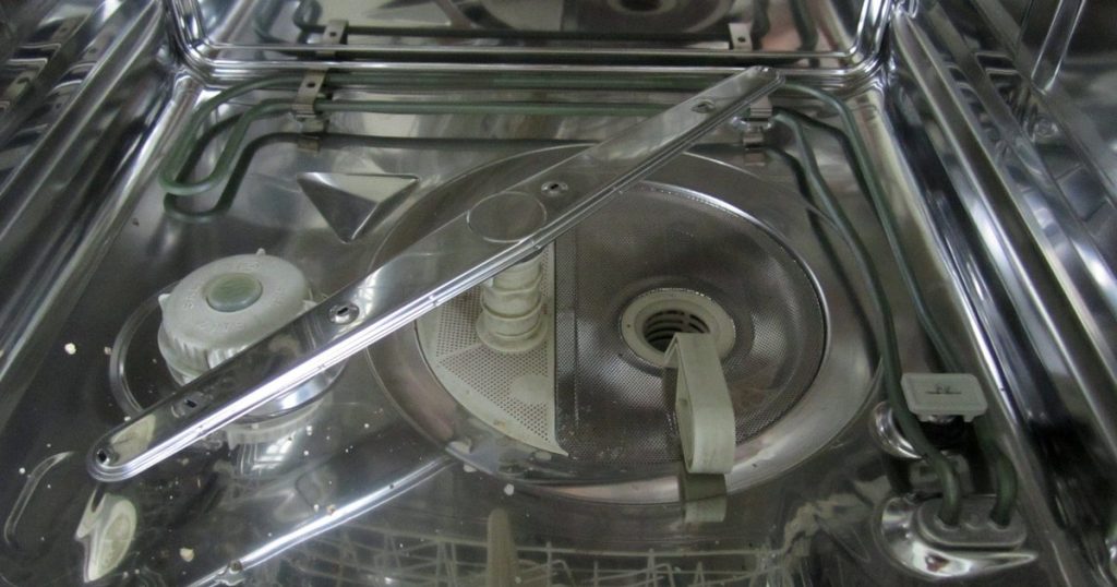 Inside of an empty dishwasher with the lower rack out, showing the spray arm, round mesh filter and drain sump in the floor of the tub