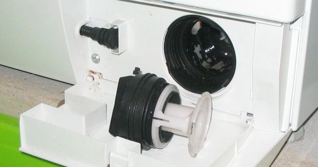 Front-load washing machine with the drain pump filter unscrewed from its housing and a green tub underneath to catch water