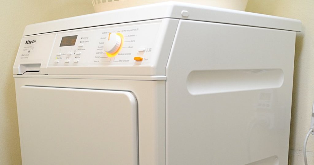 White Miele heat pump clothes dryer with its program dial lit orange and a laundry basket on top